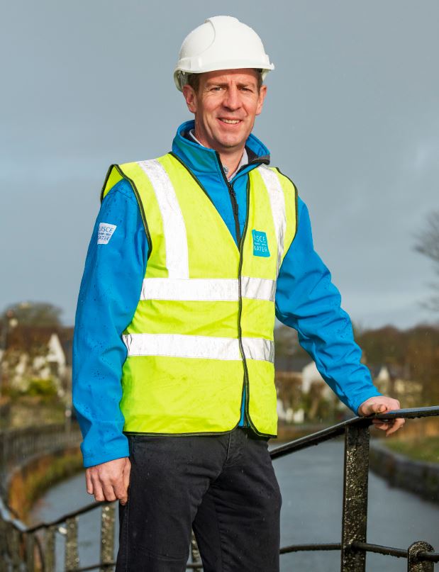 Man wearing a hi-vis jacket and white helmet standing by a railing in front of a river