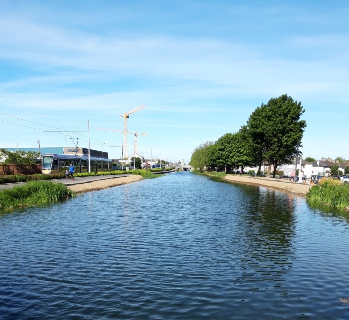 A canal by a tram line and a town with cranes in the background