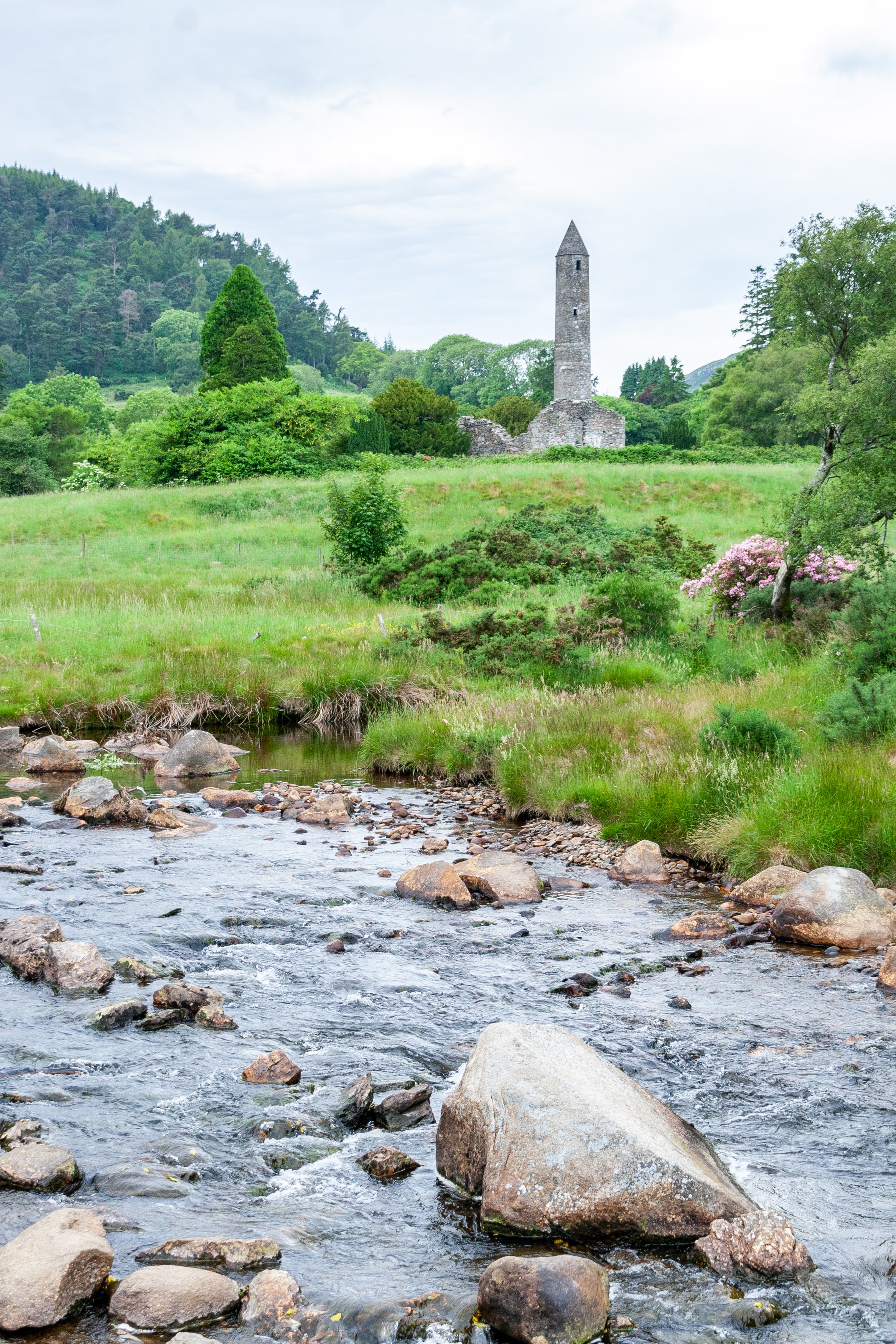 A river by a grassy field with a round tower and ruin and mountains in the distance