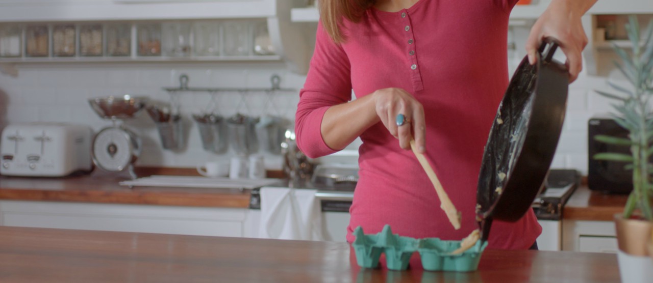 A woman in pink shirt holding a cake pan and a wooden spoon in a kitchen